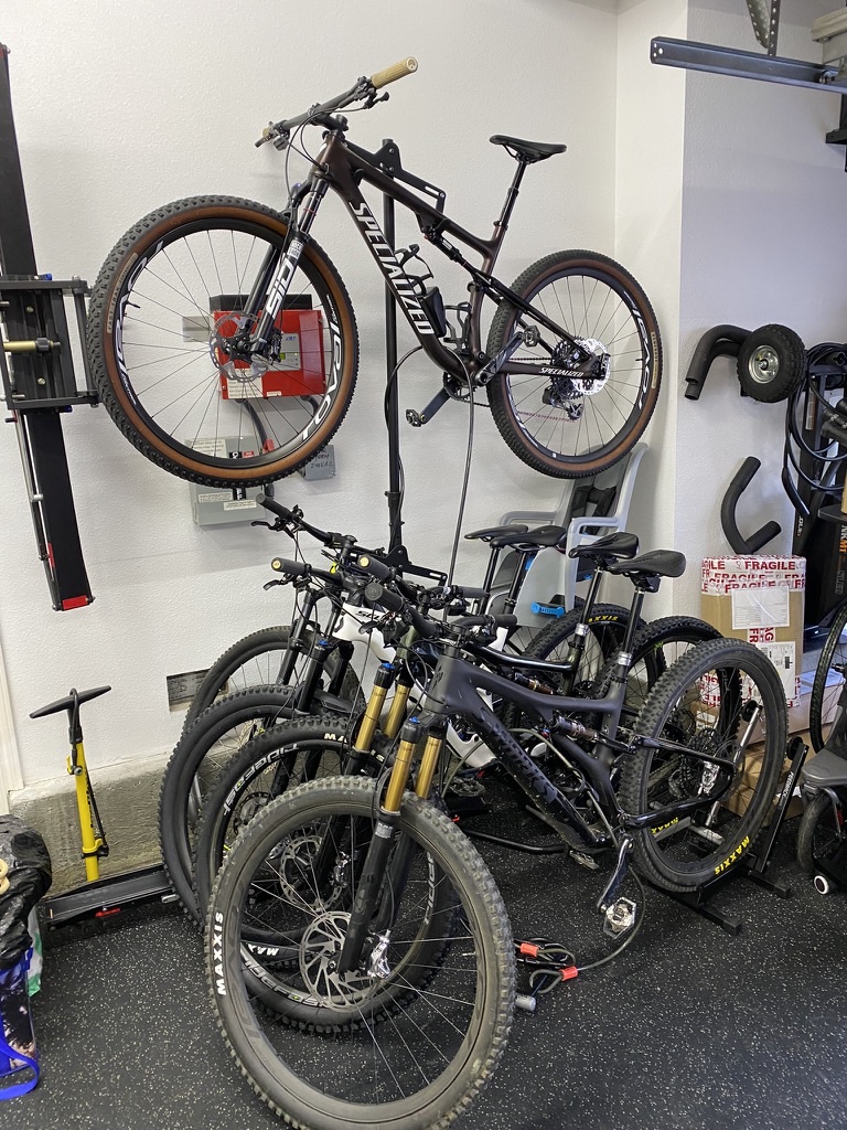 Six mountain bikes in a home garage, a Specialized full-suspension hanging on the wall above five more in floor stands, with a folded hitch rack on the wall beside them