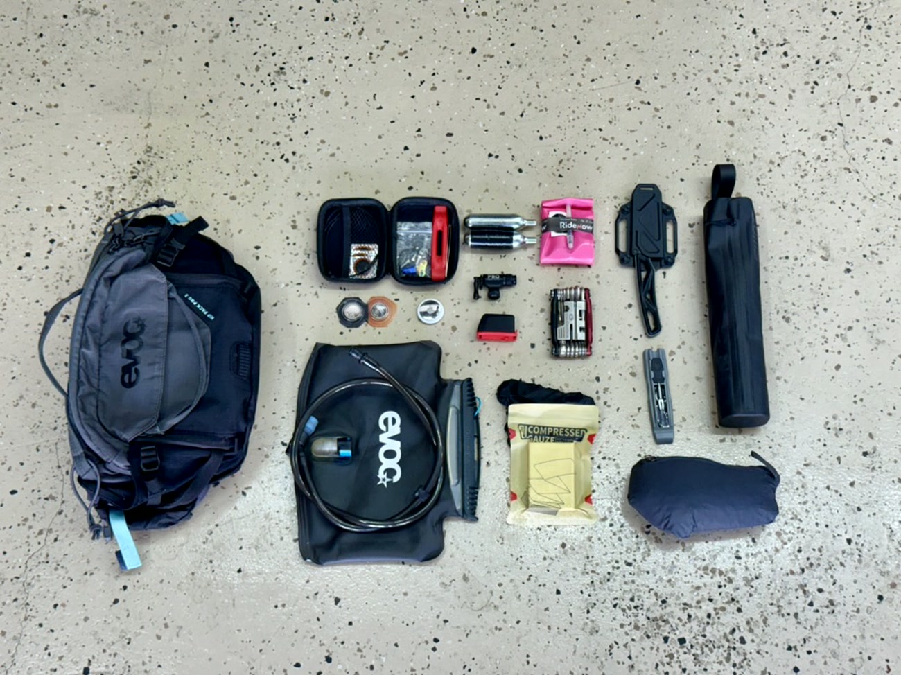 Flat-lay of a full mountain bike ride kit: EVOC hip pack and hydration bladder, repair pouch with patches and bolts, CO2 cartridges with electrical tape, multitool, tire plugs, compressed gauze, a fixed-blade knife and a packable jacket